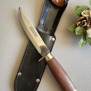 Peut inclure: Un couteau argenté avec un manche en bois brun et un fourreau en cuir noir. Le couteau porte l'inscription "Baron Nidi" gravée sur la lame.