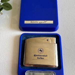 May include: A vintage silver-toned lighter with the text "Continental Reifen" in a blue case. The case is open, revealing the lighter and a velvet lining. The lid of the case has the text "Rowenta Snip".