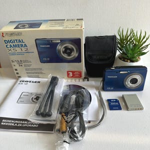 Op de afbeelding: Een blauwe Traveler XS 12 digitale camera met accessoires, waaronder een hoesje, statief, kabels en een 512MB SD-geheugenkaart. De cameradoos is zichtbaar met het model en specificaties.