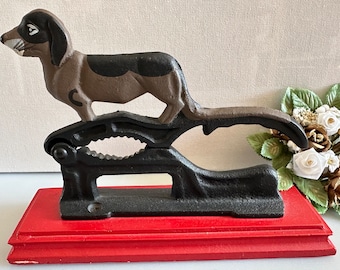 Puede incluir: Un cascanueces de hierro fundido con forma de dachshund con un acabado pintado en negro y marrón. El cascanueces está montado sobre una base de madera roja.