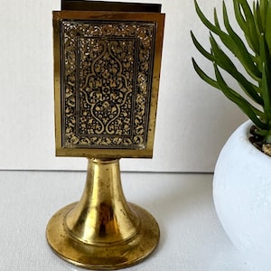 Op de afbeelding: Een gouden metalen luciferdooshouder met een ingewikkeld bloemmotief. De houder heeft een vierkante basis en een ronde voet.