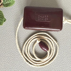 May include: A rectangular, dark maroon Sharp Shaver case with a glossy finish. A long, cream-coloured braided cord is attached to the case, featuring a matching maroon, oval-shaped bead at the end of the cord, which is coiled in a circular pattern.