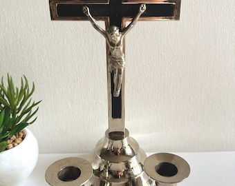 Puede incluir: Un crucifijo plateado con una figura de Jesucristo, sobre una base escalonada, acompañado de dos candelabros a juego. La cruz tiene un centro negro y un borde metálico. La inscripción INRI es visible.
