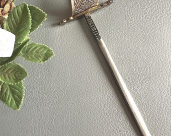 May include: A decorative hair pin with a sword-like design. The pin features a detailed, ornate hilt with engraved patterns and a long, slender, silver-colored shaft. The hilt has a gold-colored finish. The pin is likely used for securing hair in place.