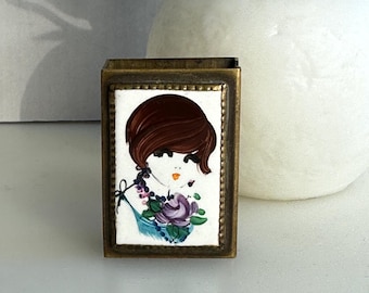 Puede incluir: Una caja de cerillas de latón vintage con una ilustración colorida de una mujer con cabello castaño y un diseño floral. La caja de cerillas tiene un borde dorado.