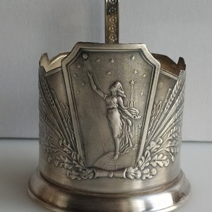 Könnte beinhalten: Silberfarbener Metall-Becherhalter mit Henkel. Die Vorderseite zeigt ein Reliefdesign einer Frau, die mit der Hand nach einem Stern greift, mit einem Globus und Weizenähren auf beiden Seiten. Der Text "СССР" ist auf der rechten Seite des Designs sichtbar.