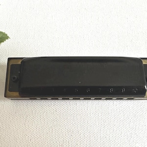 May include: A black harmonica with gold-coloured accents. The harmonica has a rectangular shape with the numbers 5 to 10 visible. The instrument is set against a white background.