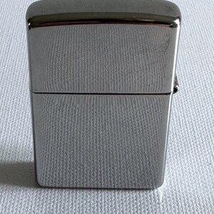 May include: A silver-colored, rectangular lighter with a hinged lid. The lighter has a smooth, reflective surface and a textured pattern on the body. The top and bottom edges are rounded. The lighter is closed in the image.