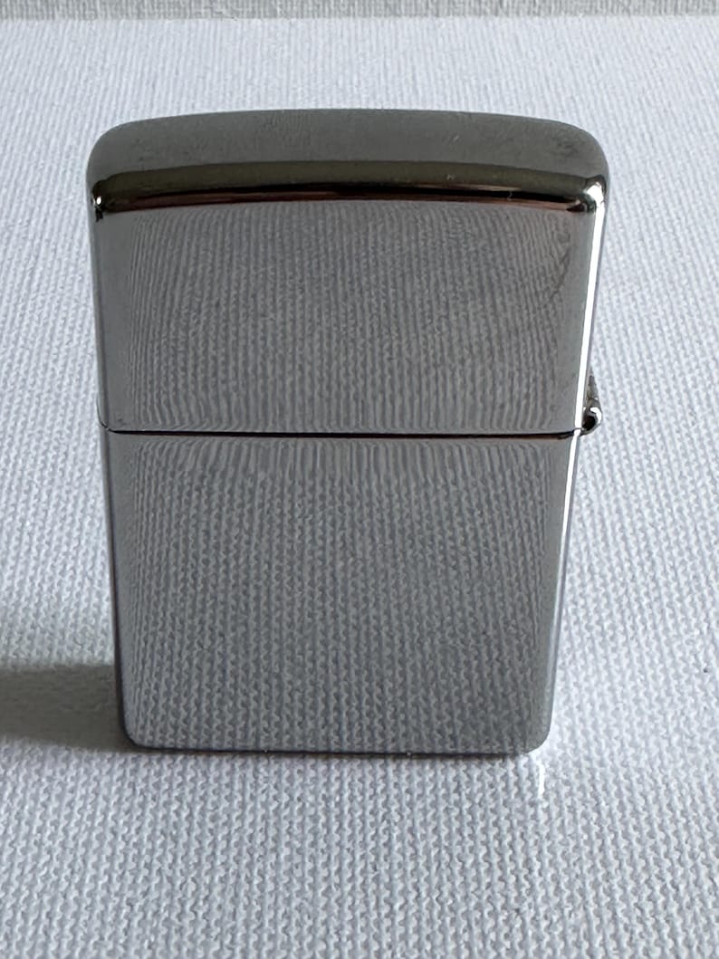 May include: A silver-colored, rectangular lighter with a hinged lid. The lighter has a smooth, reflective surface and a textured pattern on the body. The top and bottom edges are rounded. The lighter is closed in the image.