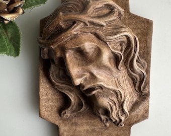 Puede incluir: Escultura en relieve marrón del rostro de un hombre, con una corona de espinas y pelo largo. La escultura está montada sobre una placa rectangular con una forma decorativa. Es probable que sea un objeto religioso.