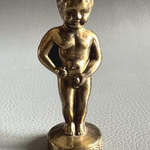 May include: A small, gold-coloured statue of a young boy urinating. The statue stands on a round base with the word "BRUXELLES" engraved. The figure has detailed facial features. A decorative item, likely a souvenir from Brussels.