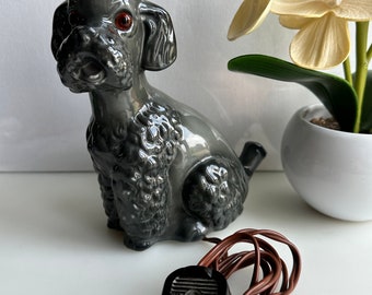 Puede incluir: Una figurita de caniche de cerámica gris con ojos rojos, un cable negro y un enchufe negro. La figurita está sentada sobre una superficie blanca.