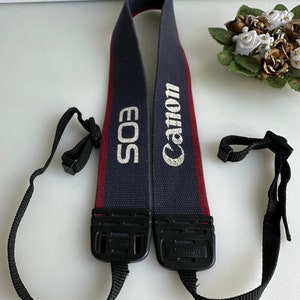 Könnte beinhalten: Ein blaues und rotes Kameraband mit dem Text "Canon EOS" in weiß.