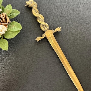 Può includere: Un tagliacarte in metallo dorato con un manico a forma di serpente attorcigliato con squame testurizzate. La guardia presenta piccole sporgenze decorative a forma di ala, e la lama è dritta e appuntita con una finitura liscia e lucida.