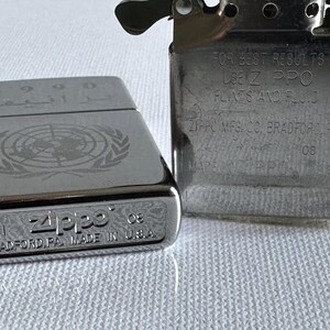 May include: Two silver Zippo lighters. One has the United Nations emblem and Arabic text. The other lighter has text that reads "FOR BEST RESULTS USE Zippo FLINTS AND FLUID" and "ZIPPO MFG. CO. BRADFORD, PA".