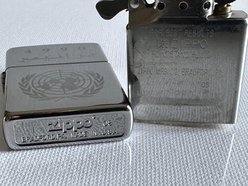 May include: Two silver Zippo lighters. One has the United Nations emblem and Arabic text. The other lighter has text that reads "FOR BEST RESULTS USE Zippo FLINTS AND FLUID" and "ZIPPO MFG. CO. BRADFORD, PA".