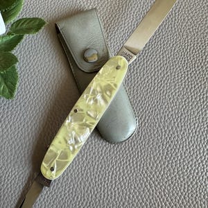 Könnte beinhalten: Ein Vintage-Nagelpflegeset mit einem Klappmesser mit einem perlmuttgelben Griff und einer silbernen Klinge. Das Set enthält ein passendes Lederetui. Das Messer ist mit dem Text "HAPO" und "Austria" gekennzeichnet.