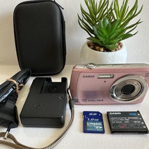 May include: A pink Casio Exilim digital camera with a 12.1-megapixel resolution, alongside a black case, charger, battery, and a 1.0 GB Kingston SD card. A small potted plant is also in the image.