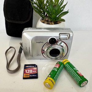 Könnte beinhalten: Eine silberne Sanyo Xacti 3,2 Megapixel Digitalkamera mit grauer Handschlaufe, schwarzer Tasche, einer 128 MB Memorex MultiMediaCard und zwei grün-gelben AA-Alkalibatterien. Das Kameraobjektiv ist mit einem roten Ring versehen.