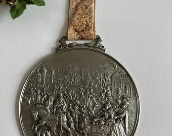 Puede incluir: Un adorno de pared circular plateado con una escena en relieve detallada de una reunión invernal, con figuras, caballos y un trineo. Tiene una parte superior decorativa y una cinta estampada para colgar. La palabra "Winter" está en la parte inferior.