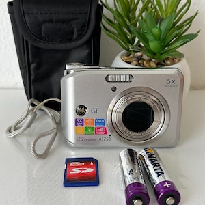 May include: Silver GE A1250 digital camera with a 5x optical zoom lens, SD card, two AA batteries, and a black carrying case. The camera has a 12.2-megapixel resolution. A small potted plant is in the background.
