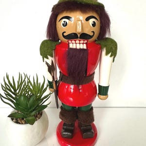 Puede incluir: Una figura de cascanueces de madera con un sombrero de fieltro verde con pluma, chaqueta roja y detalles de piel marrón. La figura tiene una barba de color púrpura oscuro, un bigote negro y sostiene una pequeña flecha marrón sobre una base roja.