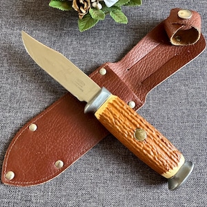 Peut inclure: Un couteau de chasse avec une lame en acier inoxydable et un manche en bois brun est rangé dans un étui en cuir brun avec une fermeture à pression métallique. La lame est marquée "SOLINGEN GERMANY".