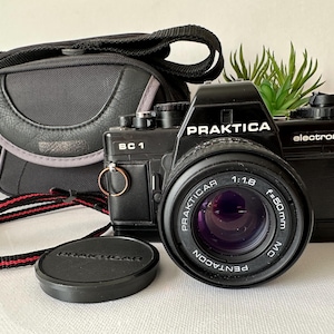 May include: A black Praktica BC 1 camera with a Pentacon lens, lens cap, and carrying case. The camera has a red and black strap. The lens is f=50mm 1:1.8. The camera is electronic.