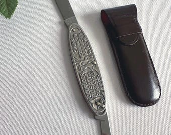 Puede incluir: Una navaja de bolsillo vintage con mango plateado y dos hojas, una extendida. El mango está grabado con texto y elementos decorativos. Una funda de cuero marrón está junto a la navaja sobre una superficie blanca.
