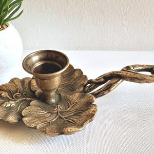 Puede incluir: Un candelero de latón vintage con diseño de hojas. El candelero tiene una copa redonda para la vela y un asa con forma de ramas entrelazadas. La base tiene forma de hoja grande. El color es dorado apagado.