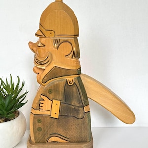 Op de afbeelding: Een houten notenkraker in de vorm van een soldaat met een punthelm. Het figuur heeft een afwerking van lichtbruin natuurhout, een donkergroen uniform met gele accenten en een lange hendel aan de achterkant om de kaak te bedienen.