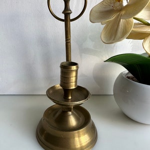 May include: A gold metal candle holder with a round base and a single candle holder on top. The candle holder has a ring on the side.