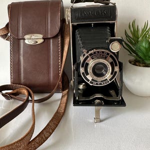 May include: Vintage Kodak camera with a brown leather case and strap. The camera is black with silver accents and features the Kodak logo. The case has a silver clasp. A small plant in a white pot is in the background.