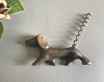 Puede incluir: Un sacacorchos vintage con forma de perro salchicha. El sacacorchos de metal tiene una cola en espiral y un cuerpo de color bronce. El perro está en posición de pie. El sacacorchos está sobre una superficie gris.