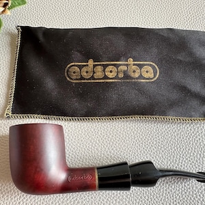 May include: A burgundy-colored smoking pipe with a black stem and mouthpiece, accompanied by a black pouch with the gold text "adsorba". The pipe has a curved design and a smooth finish, suggesting a classic style.