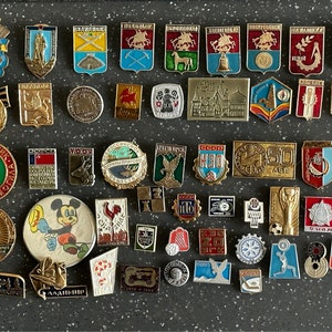 Peut inclure: Une collection d'épingles en émail vintage avec une variété de motifs, y compris des animaux, des bâtiments et des motifs abstraits. Les épingles sont principalement rouges, bleues et vertes, avec quelques accents dorés. Les épingles sont disposées en rangées sur une surface gris foncé.