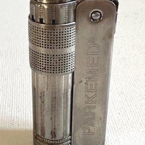 Puede incluir: Un encendedor plateado vintage con cuerpo rectangular y tapa abisagrada. El encendedor tiene un patrón texturizado, similar a una rejilla, alrededor del centro. El texto "PARKEMED" está grabado en el lateral.