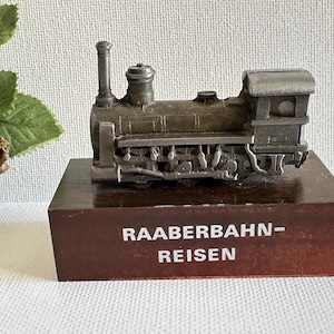 May include: A detailed miniature train engine made of metal, mounted on a dark brown wooden base. The base has the text "RAABERBAHN-REISEN" in white. The train features a smokestack and intricate detailing.