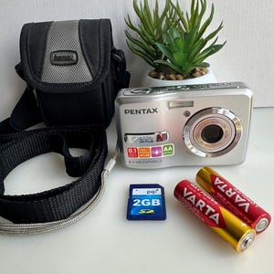 Puede incluir: Cámara digital Pentax plateada con correa y funda de transporte negras. Incluye una tarjeta SD de 2 GB y dos pilas AA Varta. También hay una pequeña planta en maceta en la imagen.