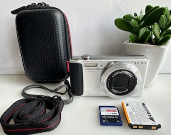Puede incluir: Una cámara digital Praktica sumedia 10-2120 plateada con un objetivo de zoom óptico 12,5x y 16,1 megapíxeles. La cámara está en una funda de transporte negra con una cremallera roja. La cámara está junto a una tarjeta de memoria y una batería.