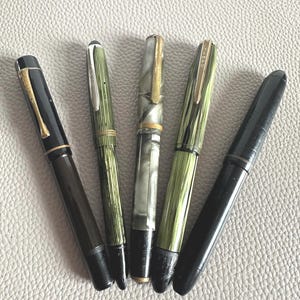 Könnte beinhalten: Fünf Vintage-Füllfederhalter. Die Stifte haben schwarze, grüne und marmorierte Schäfte mit goldenen und silbernen Akzenten. Die Stifte sind auf einer strukturierten, hellgrauen Oberfläche angeordnet.