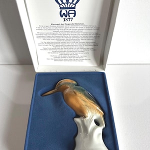 Puede incluir: Figurita de porcelana de un martín pescador, presentada en una caja. El ave presenta tonos naranja, azul y verde. La caja incluye texto en alemán y el logotipo de la marca "WA 1877".
