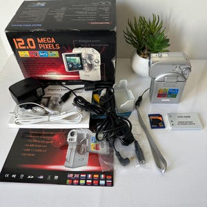 Puede incluir: Videocámara digital plateada con una caja que dice "12.0 MEGA PIXELS". El kit incluye un cargador, cables, tarjetas de memoria y una batería. La caja también enumera características como zoom digital 8x y un micrófono incorporado.