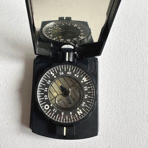 May include: Black compass with a hinged lid, open to reveal the compass dial and a mirrored surface. The dial features degree markings and cardinal directions. The compass is designed for navigation and outdoor use.