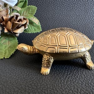Könnte beinhalten: Eine goldfarbene Metall-Schildkrötenfigur mit detailliertem Panzer und Beinen. Die Schildkröte befindet sich auf einer strukturierten schwarzen Oberfläche, mit künstlichen Blumen im Hintergrund, darunter weiße, goldene und grüne Elemente.