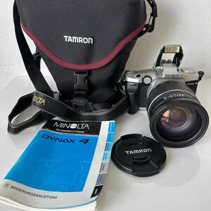 May include: A black camera bag with a red trim and the brand name "Tamron" is next to a Minolta Dynax 4 camera with a lens and a lens cap. A manual is also present.