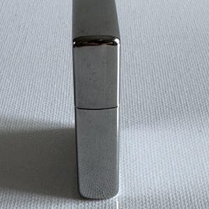 May include: A silver-colored, rectangular lighter with a hinged lid. The lighter has a smooth, reflective surface and a classic design. The lighter is shown against a white background.
