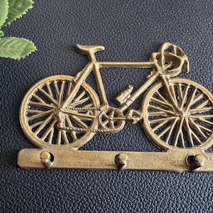 Op de afbeelding: Een messingkleurige fietssleutelhouder met drie haken. De fiets heeft gedetailleerde spaken, een waterfles en een helm. De sleutelhouder is gemonteerd op een donkerblauwe ondergrond.