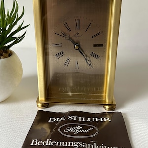 Puede incluir: Un reloj de viaje Royal dorado con una esfera rectangular y números romanos. El reloj tiene un asa y descansa sobre pequeños pies. Un manual de instrucciones marrón está delante del reloj, con el texto "DIE STILUHR" y "Royal".
