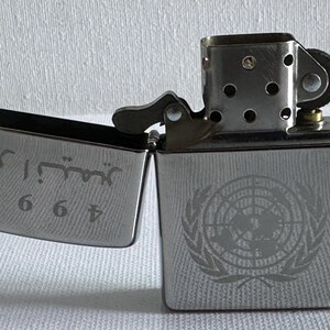 May include: A silver-colored lighter with the lid open, revealing engraved text and numbers. The body of the lighter features a detailed emblem. The lighter is a classic design, suitable for various uses.